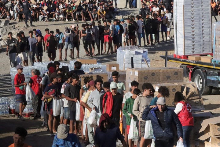 Una fila de migrantes espera recibir alimentos que entrega la Cruz Roja en la playa del Trampolín, este jueves en Ceuta. Foto: EFE 